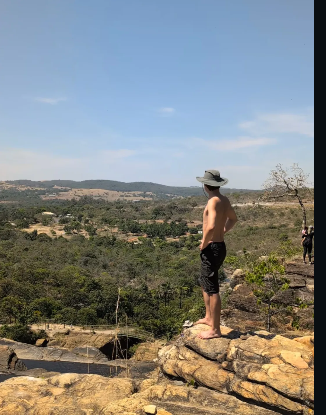 Félmeztelen kalapos fiatal férfi áll a természetben egy sziklán a napsütésben és csodálja a tájat